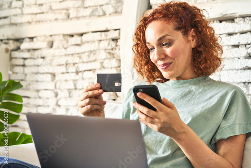 Fotografie, Tablou Young redhair woman sitting on her sofa doing online shopping on laptop in morni