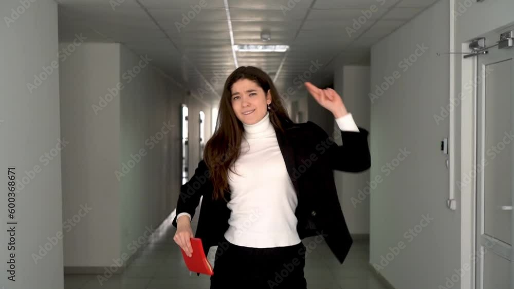 Enthusiastic young brunette in smart clothing with red notebook dancing expressively in the middle of empty grey lit hallway of office building Enthusiastic young brunette in smart clothing with red notebook dancing expressively in the middle of empty grey lit hallway of office building