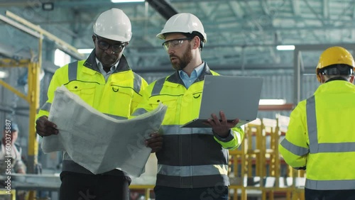 Two men move and communicate with each other on construction site. Engineers dressed in yellow vests and helmets. African American architect holding blueprints. Worker is gesturing and holding laptop.