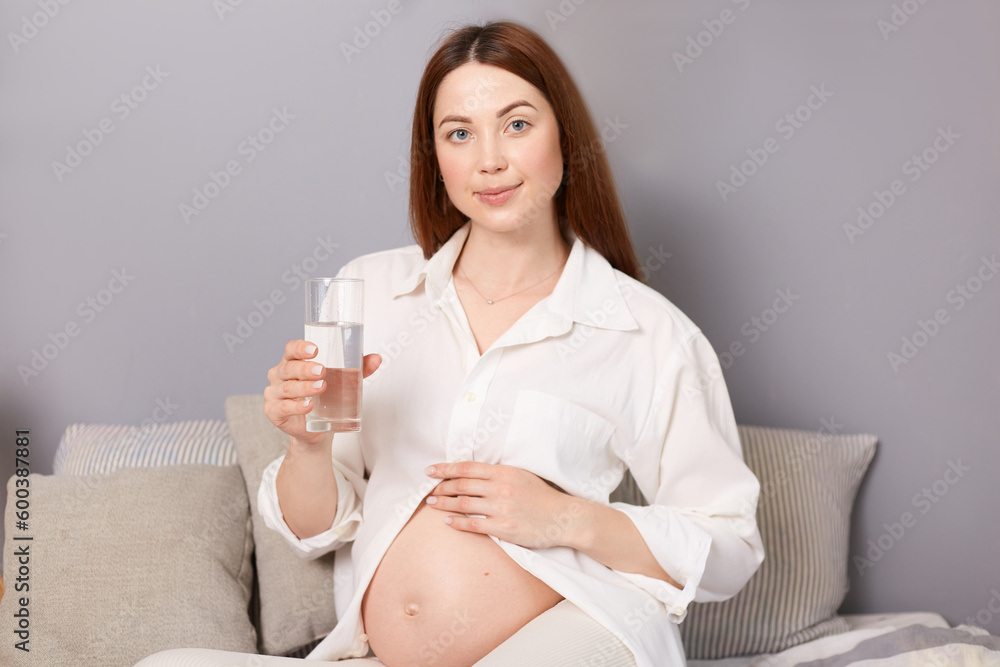 Smiling young pregnant woman in white shirt and leggings is sitting on a bed, enjoying a fresh beverage and touching her bare belly while drinking water. Smiling young pregnant woman in white shirt and leggings is sitting on a bed, enjoying a fresh beverage and touching her bare belly while drinking water.