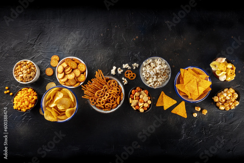 Salty snacks, party mix. An assortment of crispy appetizers, shot from above on a black background with copy space. Potato and tortilla chips, crackers, popcorn etc