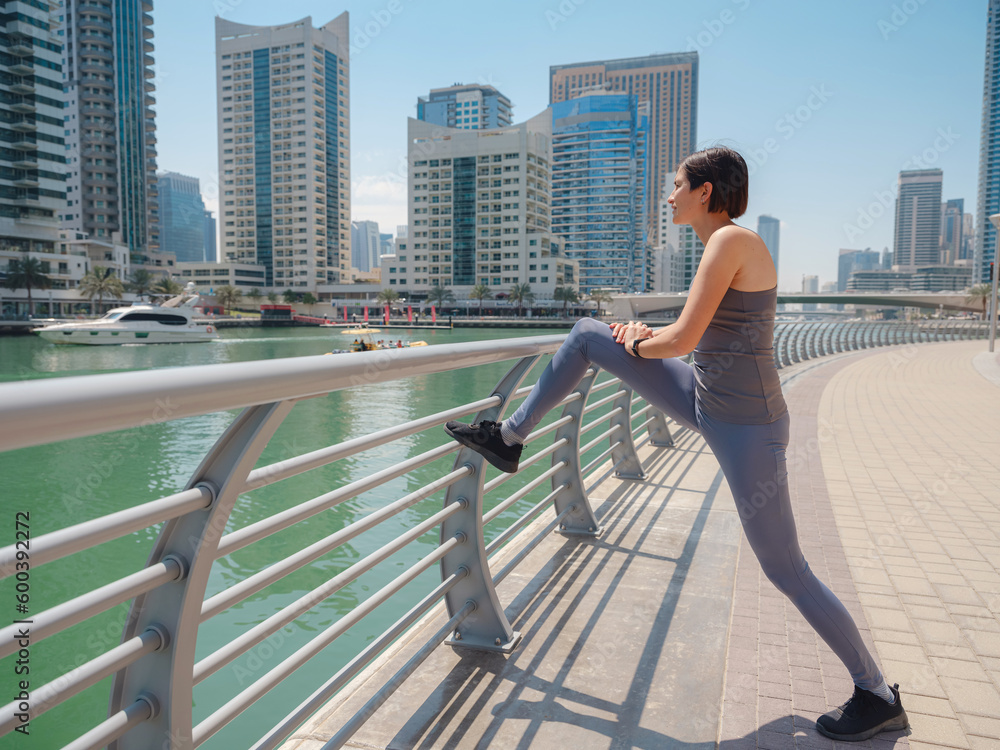 Smiling asian woman stretching legs over urban skyline in Dubai marina. woman enjoying daily routine warming up before running at morning. Sporty lady doing leg stretches before workout