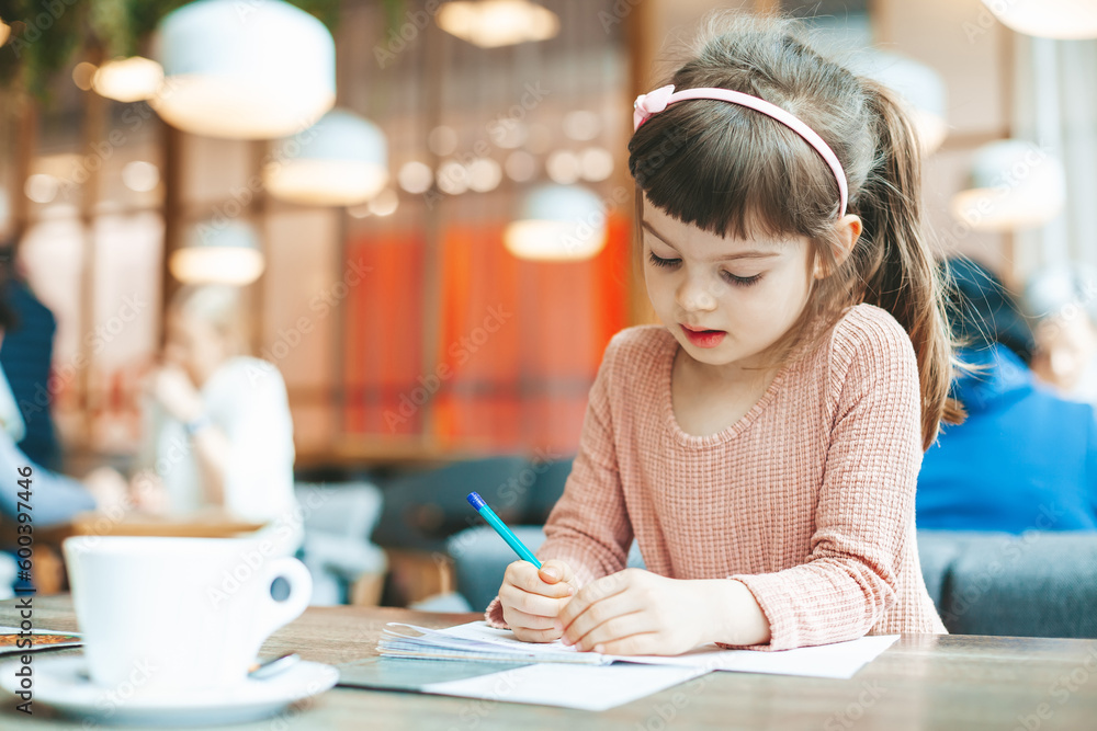 Cute little girl writing something in a notebook sitting in a cafe Cute little girl writing something in a notebook sitting in a cafe