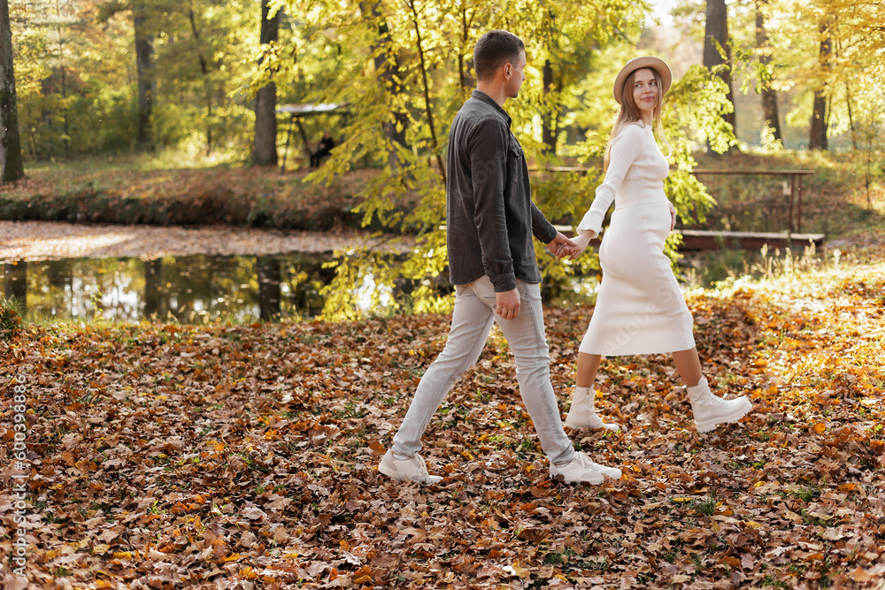 Fototapeta premium couple, man and pregnant wife are holding hands and walking in the autumn park on sunset. Happy family enjoying pregnancy on nature. The concept of Mother's Day and Women's Day.