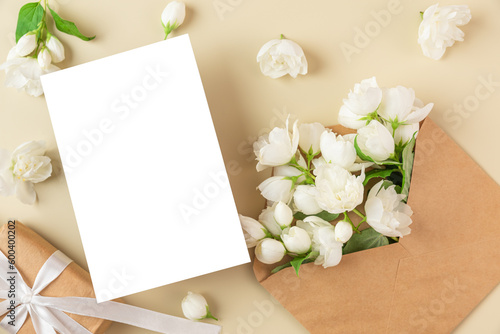 Blank greeting card with white jasmine flowers and gift box on beige background. Wedding invitation. Mock up. Flat lay