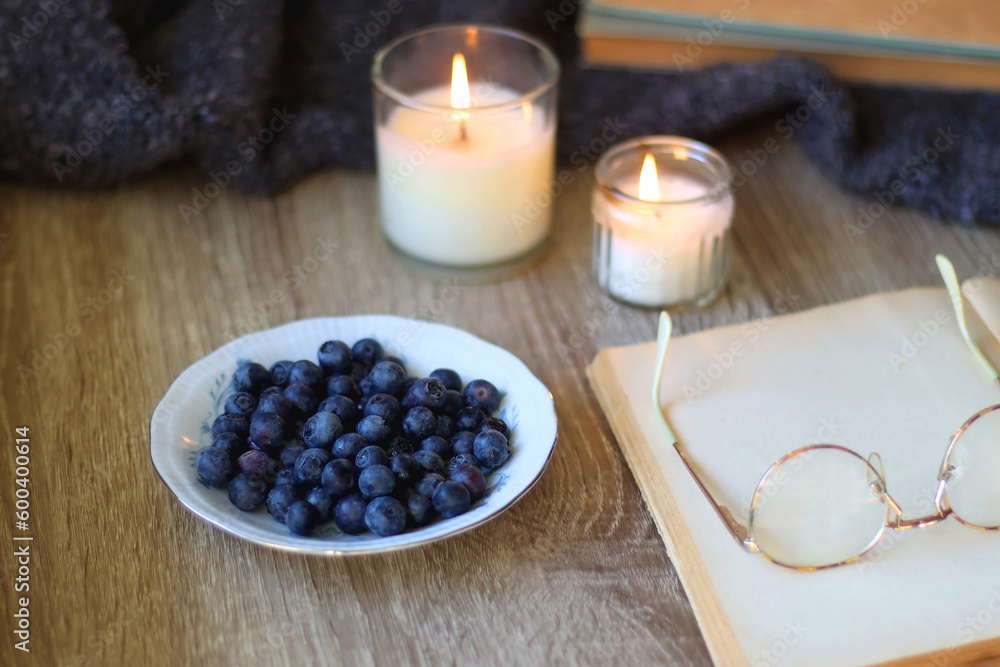 Cup of blueberries, set of books, phone, eyeglasses, lit candles and blanket on the table. Hygge at home. Selective focus. Cup of blueberries, set of books, phone, eyeglasses, lit candles and blanket on the table. Hygge at home. Selective focus.