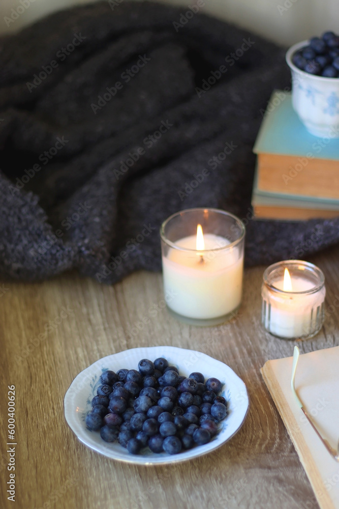 Cup of blueberries, set of books, phone, eyeglasses, lit candles and blanket on the table. Hygge at home. Selective focus. Cup of blueberries, set of books, phone, eyeglasses, lit candles and blanket on the table. Hygge at home. Selective focus.