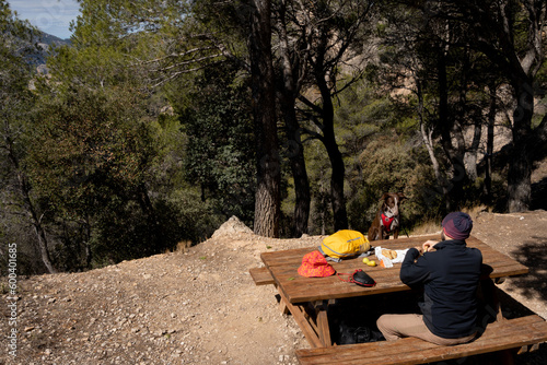 man with dog eating some sausage on a picnic table, dressed as a mountaineer with a headscarf, yellow backpack and flower cap on the table