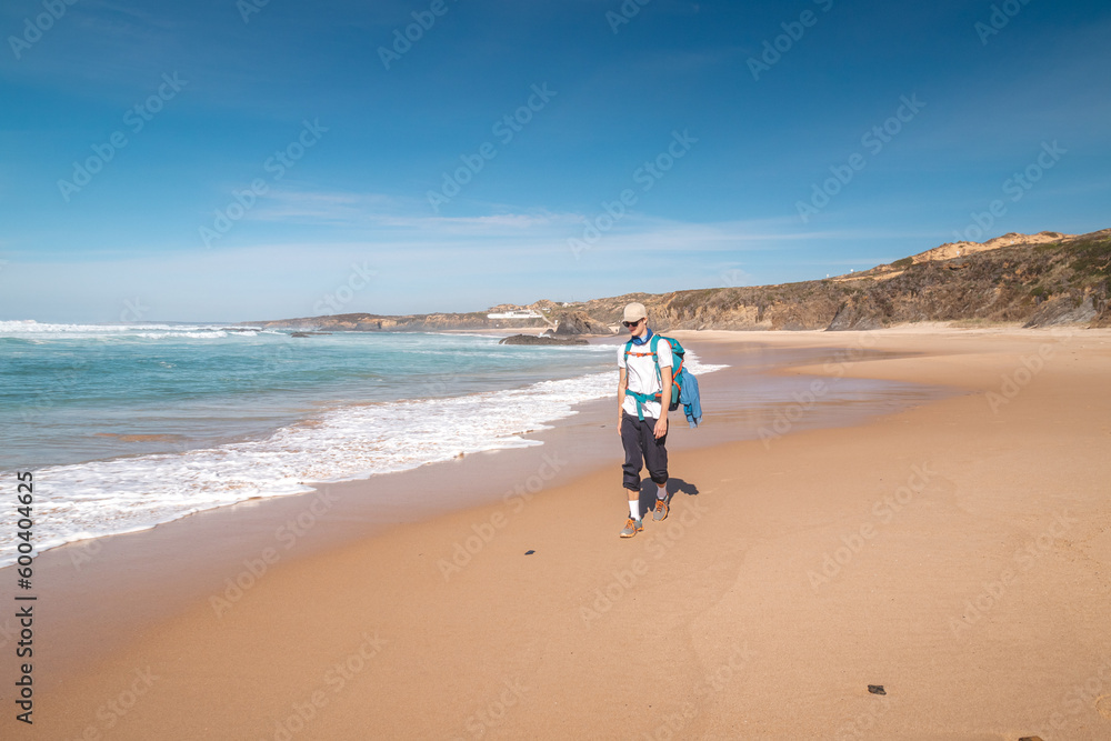 Backpacker walks along Praia do Almograve with a smile on his face. The joy of moving and discovering new places. Odemira region, western Portugal. Wandering along the Fisherman Trail, Rota Vicentina