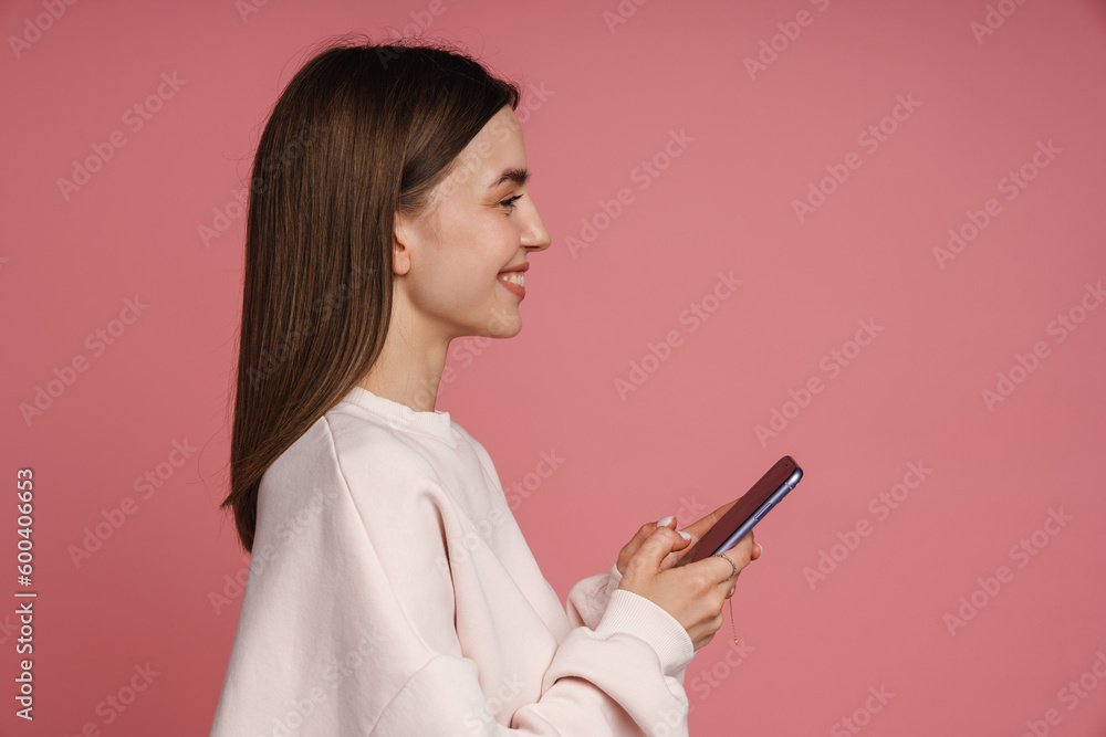 Profile of smiling woman using mobile phone while standing isolated over pink background
