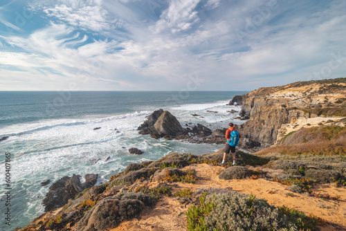 Joyful expression of a young backpacker standing on the edge of a cliff overlooking the Atlantic Ocean in the Odemira region, western Portugal. Wandering along the Fisherman Trail, Rota Vicentina