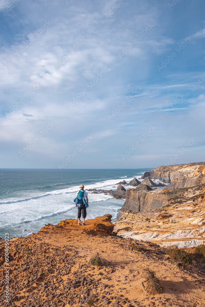 Joyful expression of a young backpacker standing on the edge of a cliff overlooking the Atlantic Ocean in the Odemira region, western Portugal. Wandering along the Fisherman Trail, Rota Vicentina