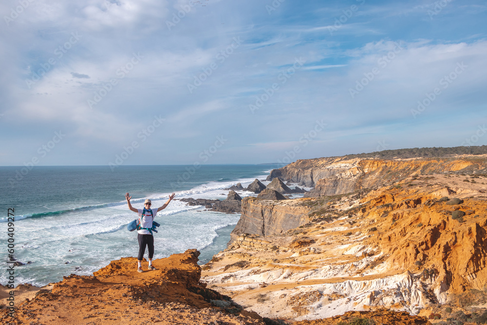 Joyful expression of a young backpacker standing on the edge of a cliff overlooking the Atlantic Ocean in the Odemira region, western Portugal. Wandering along the Fisherman Trail, Rota Vicentina
