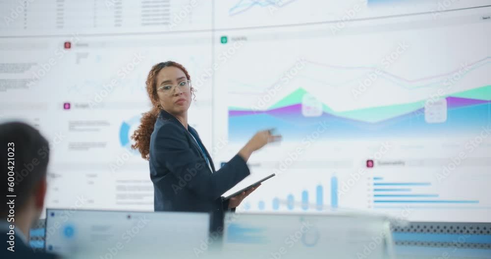 Hispanic Female Senior Data Scientist Reviewing Reports Of Risk Management Department On Big Digital Screen In Monitoring Room. Diverse Consulting Company Employees Working Behind Desktop Computers. Hispanic Female Senior Data Scientist Reviewing Reports Of Risk Management Department On Big Digital Screen In Monitoring Room. Diverse Consulting Company Employees Working Behind Desktop Computers.