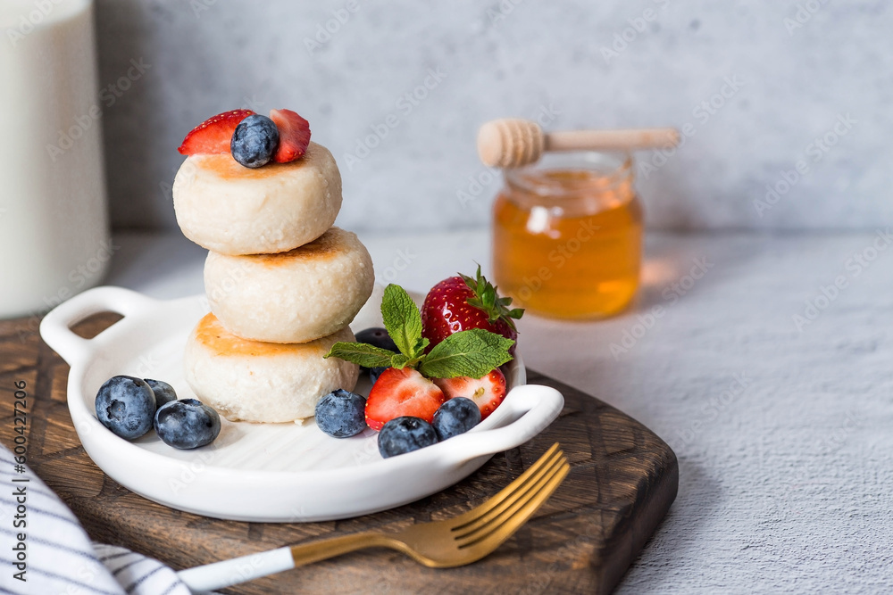 Obraz premium Cottage cheese pancakes, Russian cheesecakes with fresh blueberries, strawberries, honey and mint leaves on a stylish wooden board on a gray concrete table. Healthy and delicious breakfast.