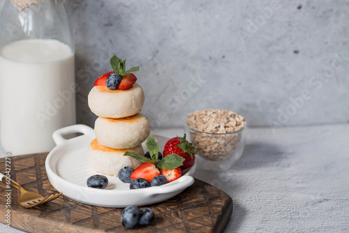 Cottage cheese pancakes, Russian cheesecakes with fresh blueberries, strawberries and mint on a stylish wooden board on a gray concrete table. Natural products. Healthy and delicious breakfast.
