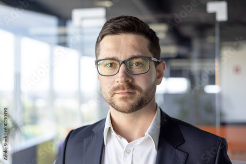 Portrait of young successful serious businessman, broker businessman close up in glasses and business suit looking at camera, workplace man working inside office, thinking about decision