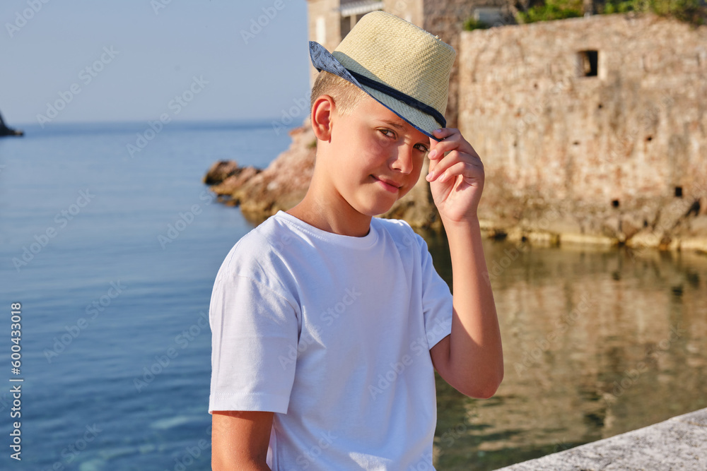 Portrait of a teenager boy in a white t-shirt and straw hat sitting near the sea.