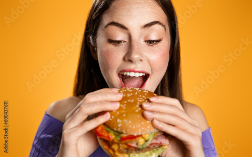 Close up of beautiful woman bites big delicious burger. Hungry girl eats hamburger with pleasure  looks with appetite at junk food  breaking diet  stands over orange background