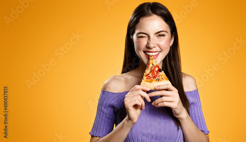 Brunette girl bites slice of pizza, smiles and winks with joy, eats takeaway food, orders delivery in restaurant app, hungry woman with junkfood against orange background