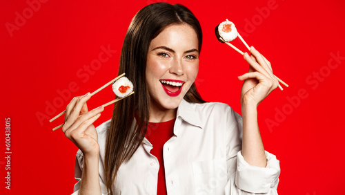 Enthusiastic woman with chopsticks, eats sushi rolls and laughs, orders delivery takeaway from asian restaurant. Girl enjoys asian food, japanese maki with salmon and tuna, red background photo