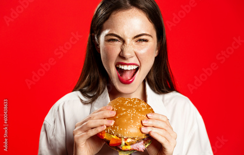 Cheerful pretty woman with a hamburger in the hands of a fast food diet  eats delicious burger on red background