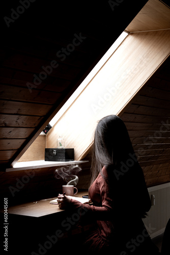 Woman with long hair writing next to a attic window drinking tea and natural light, writer working