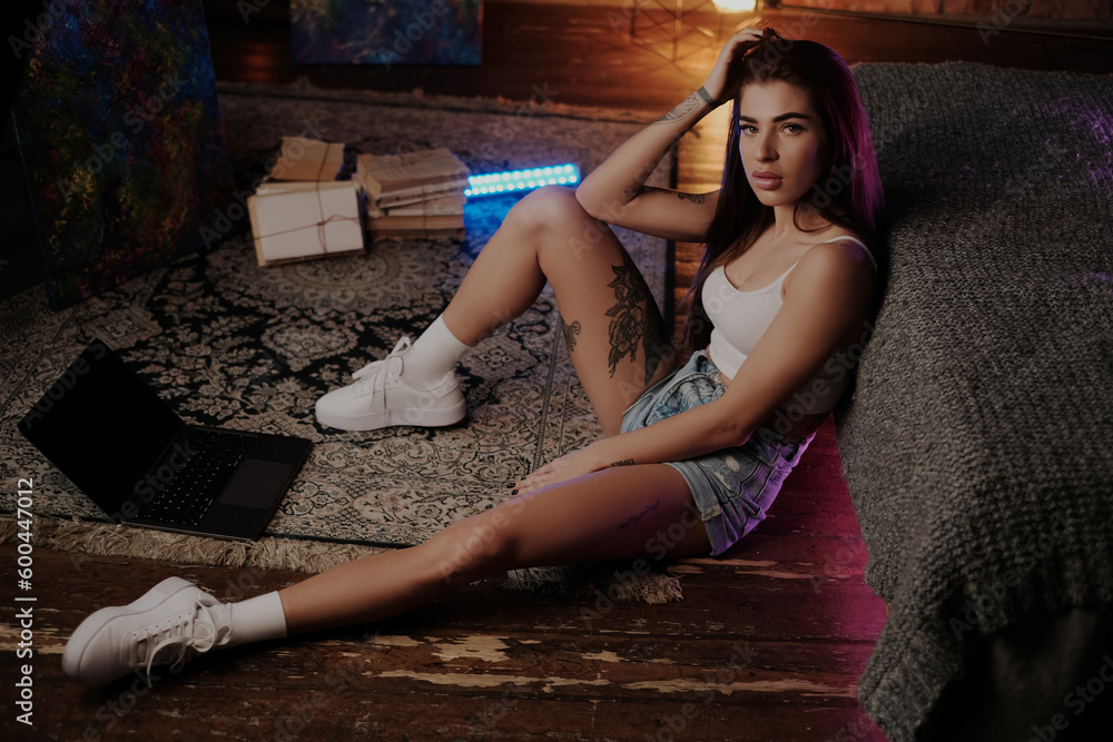 Fototapeta premium Seductive female model with tattoos and laptop sits on floor looking at camera around neon lightning in living room.