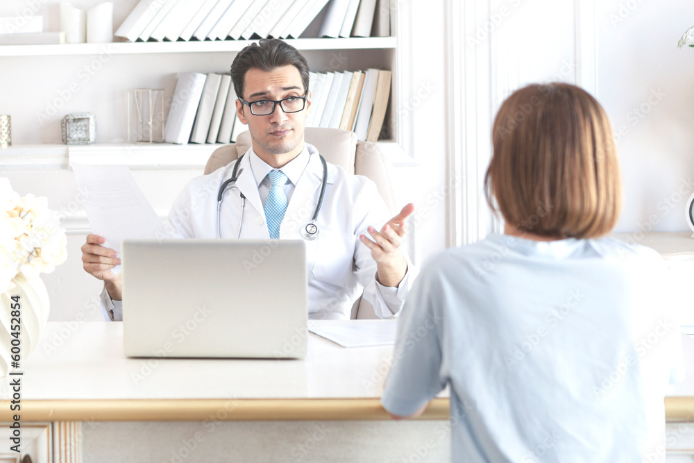 Young handsome physician in white medical gown receiving female patient in his modern workplace Young handsome physician in white medical gown receiving female patient in his modern workplace