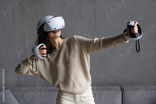 A woman in virtual reality glasses with joysticks in her hands, sitting on a couch and traveling through a meta universe. VR helmet for entertainment, learning and gaming.