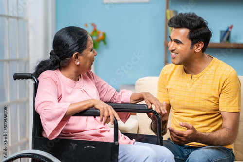 Happy indian adult son with disabled senior mother on wheelchair spending time by talking at home - concept of family support, home carea and motherhood