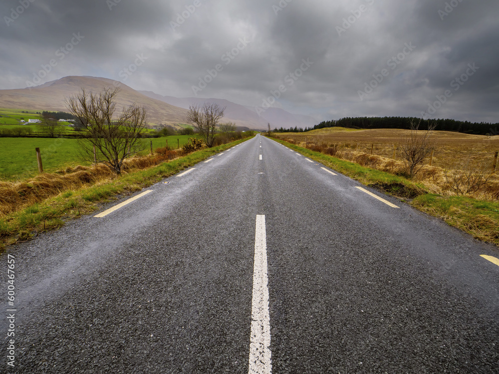 Small narrow asphalt road to mountains in low cloudy sky. Travel background. Calm Irish nature landscape. Transportation and tourism concept. West of Ireland.