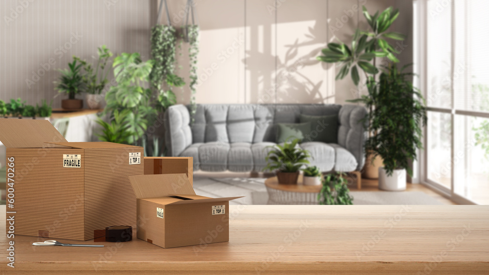 Wooden table, desk or shelf with stack of cardboard boxes over blurred view of kitchen, living and dining room, houseplants interior design, moving house concept with copy space Wooden table, desk or shelf with stack of cardboard boxes over blurred view of kitchen, living and dining room, houseplants interior design, moving house concept with copy space