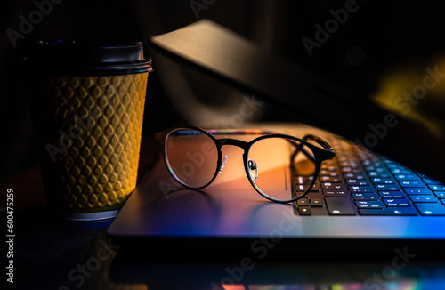 A laptop half closed in the dark with colourful glow, glasses and paper cup. photo