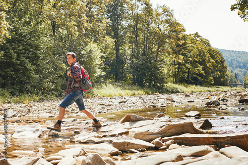 Trekking with backpack concept image. Backpacker wearing trekking boots crossing mountain river. Man hiking in mountains during summer trip