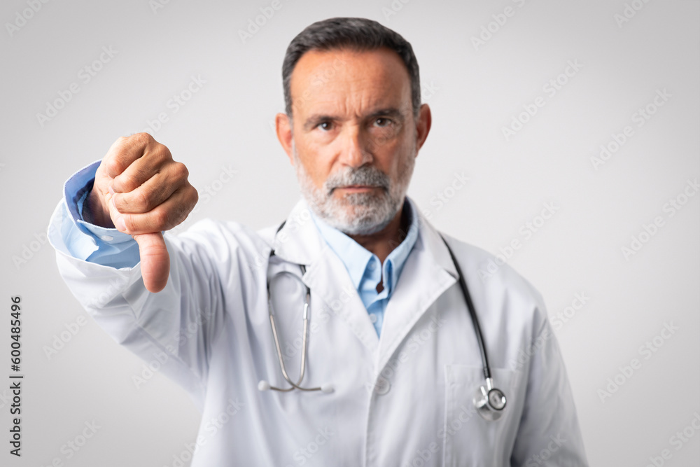 Serious confident european old man doctor in white coat with stethoscope show thumb down sign Serious confident european old man doctor in white coat with stethoscope show thumb down sign