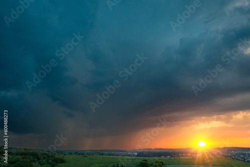 Gorgeous sunset view of a rainy dark cloud over summer country hills. The shining sun is low over the horizon—dramatic scenery.