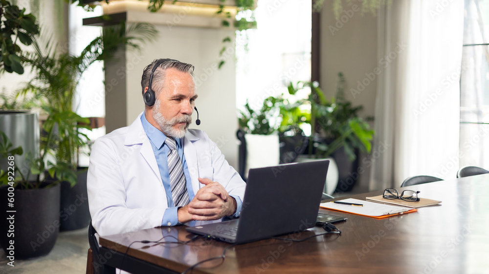 Fototapeta premium Senior caucasian doctor online video call conference consult patient with headset on laptop computer. Doctor online consultation and telehealth medicine