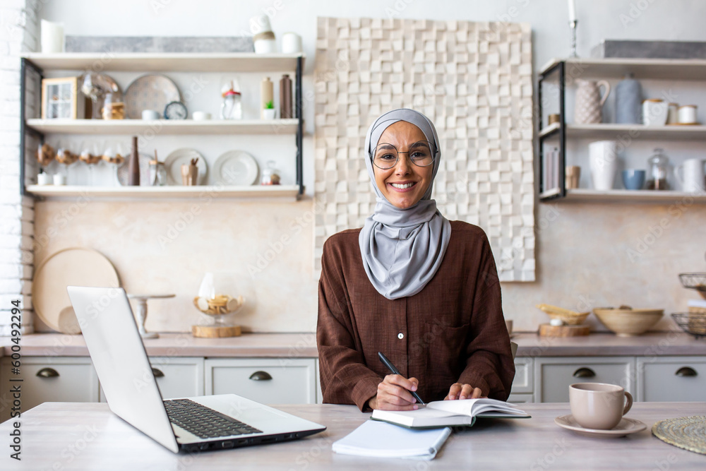 Portrait of a young female student of oriental origin in a hijab sitting at home in the kitchen and studying remotely from a laptop. Notes lectures, lessons. Smiling, she looks at the camera. Portrait of a young female student of oriental origin in a hijab sitting at home in the kitchen and studying remotely from a laptop. Notes lectures, lessons. Smiling, she looks at the camera.