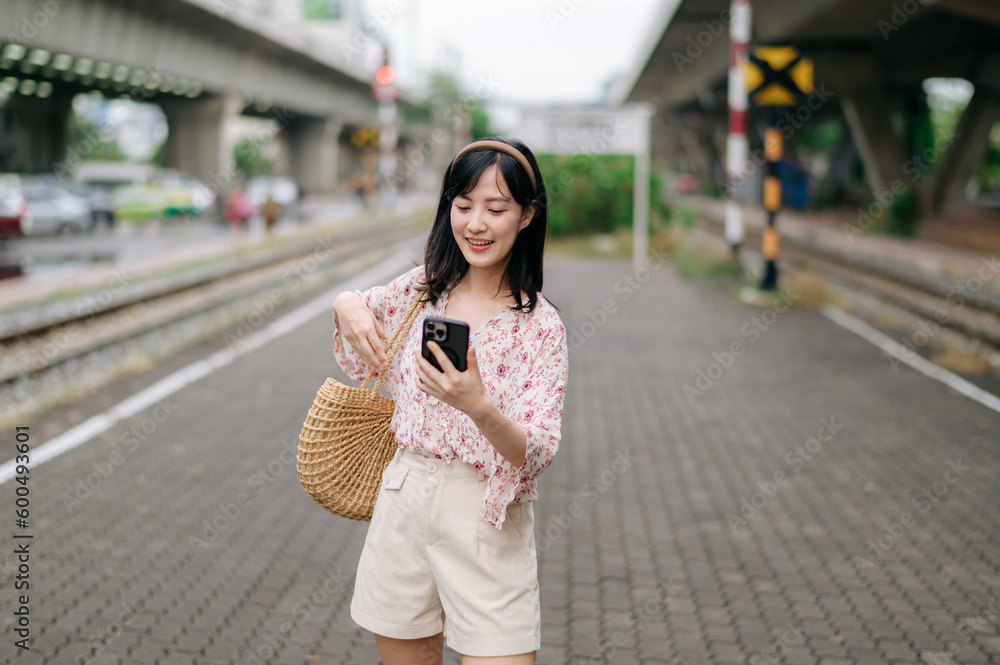 Fototapeta premium Asian young woman traveler with weaving basket using a mobile phone beside railway train station in Bangkok. Journey trip lifestyle, world travel explorer or Asia summer tourism concept.