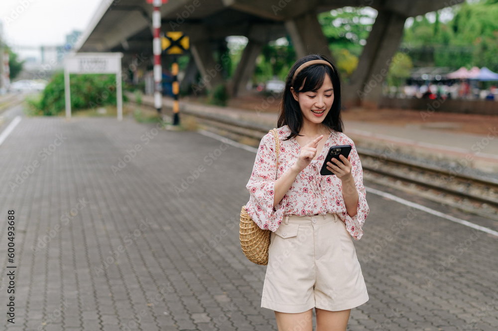 Fototapeta premium Asian young woman traveler with weaving basket using a mobile phone beside railway train station in Bangkok. Journey trip lifestyle, world travel explorer or Asia summer tourism concept.
