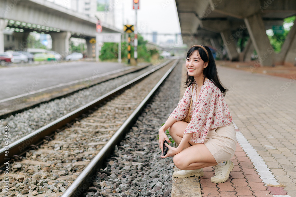 Fototapeta premium Asian young woman traveler with weaving basket using a mobile phone beside railway train station in Bangkok. Journey trip lifestyle, world travel explorer or Asia summer tourism concept.