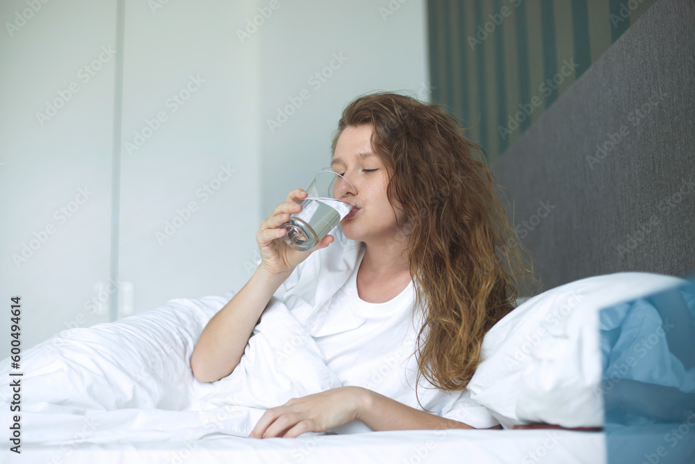 Young woman in the bed in bedroom at home in the morning lying under white blanket and drinking pure fresh water from glass Young woman in the bed in bedroom at home in the morning lying under white blanket and drinking pure fresh water from glass