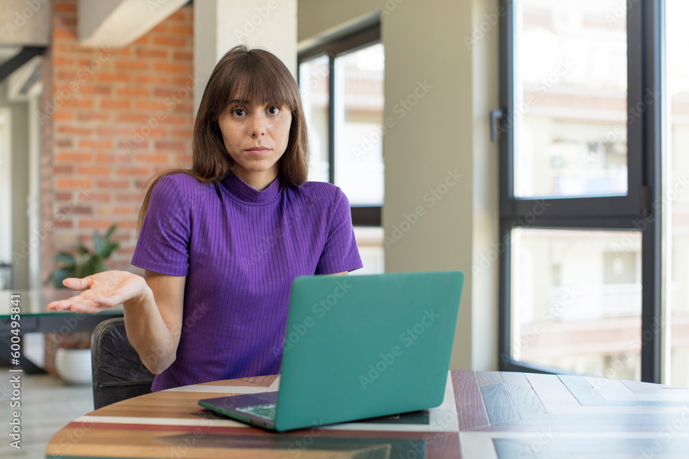 young pretty woman shrugging, feeling confused and uncertain. desk laptop concept young pretty woman shrugging, feeling confused and uncertain. desk laptop concept