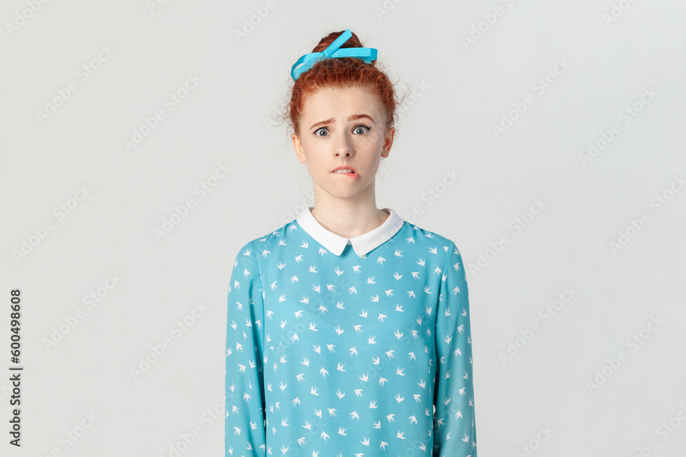 Portrait of scared frighten ginger woman with bun hairstyle, looking at camera and biting her lips, being nervous, wearing blue dress. Indoor studio shot isolated on gray background.