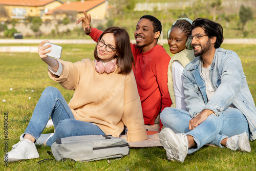 Glad multiethnic millennial students study, sit on grass, taking selfie together to blog at university
