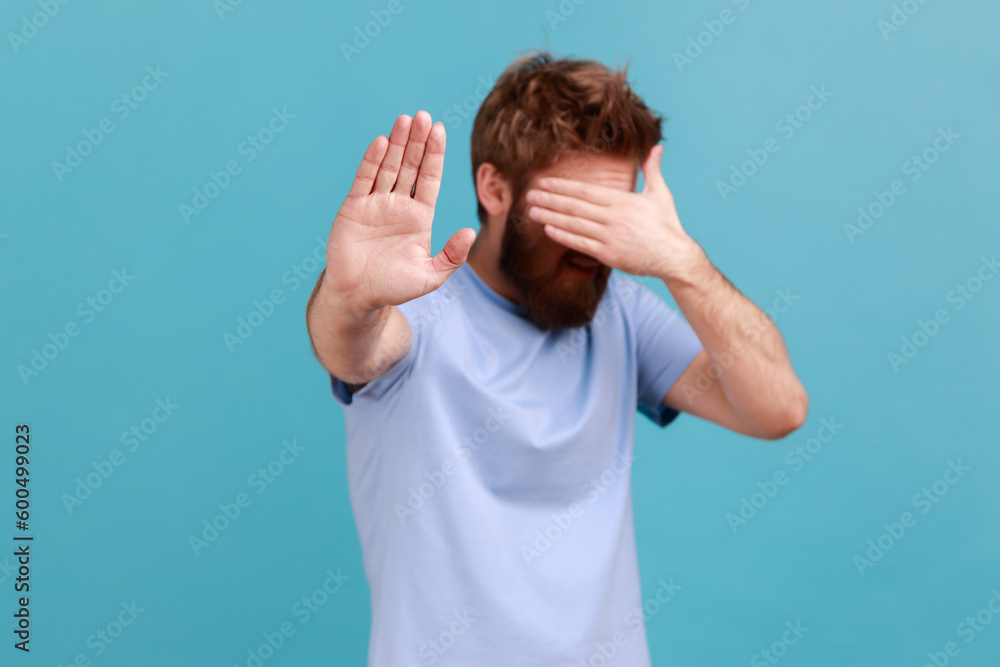 © khosrork - Portrait of bearded man standing, closed his eyes with hands and showing stop gesture, feeling stressed afraid, refusing to watch scary content. Indoor studio shot isolated on blue background.