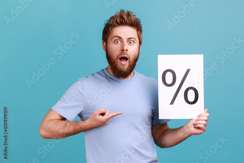 Portrait of bearded man holding paper with percent sign inscription, looking at camera with open mouth and shocked expression. Indoor studio shot isolated on blue background. © khosrork