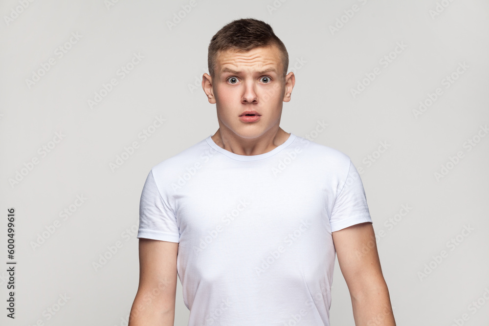 Fototapeta premium Portrait of astonished surprised attractive teenager boy wearing T-shirt standing looking at camera with big eyes, being frighten and scared. Indoor studio shot isolated on gray background.