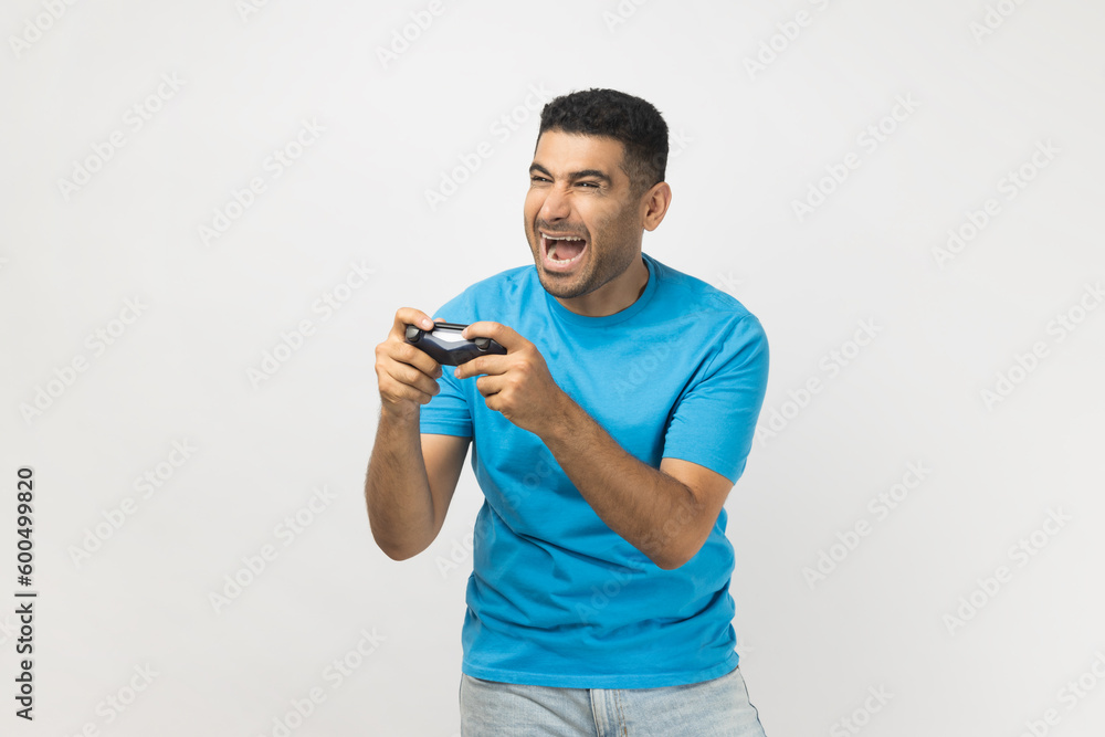 Portrait of excited extremely happy unshaven man gamer wearing blue T- shirt standing playing video games, holding joystick in hands. Indoor studio shot isolated on gray background. Portrait of excited extremely happy unshaven man gamer wearing blue T- shirt standing playing video games, holding joystick in hands. Indoor studio shot isolated on gray background.